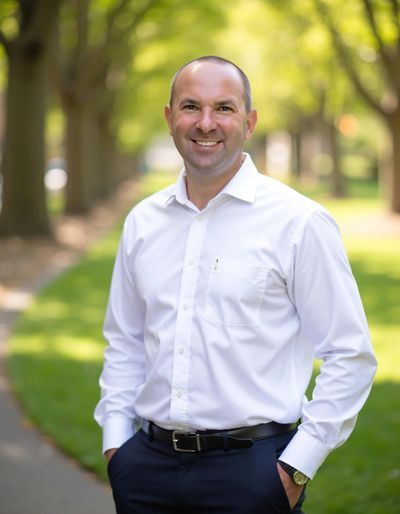 Smiling man in white shirt standing on a tree-lined pathway.
